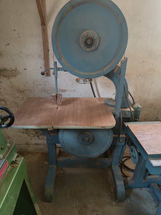 Gammaldags blå bandsåg från 50-talet med träbord och stålstativ, belägen inomhus. Drivs med 380 V elmotor. Finns i ett snickeri.