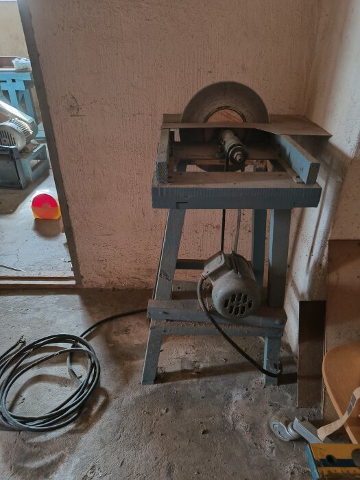 Gammal cirkelsåg på trästativ med kilremsdrift och 380 V motor, placerad i ett verkstadsutrymme med elkabel vid sidan.