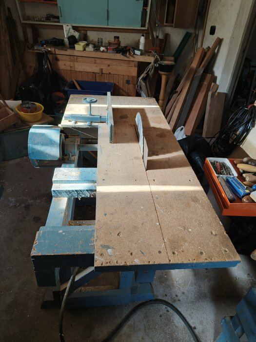 Gammal snickerimaskin från 50-talet på trästativ i en verkstad, omgiven av verktyg och träplankor.