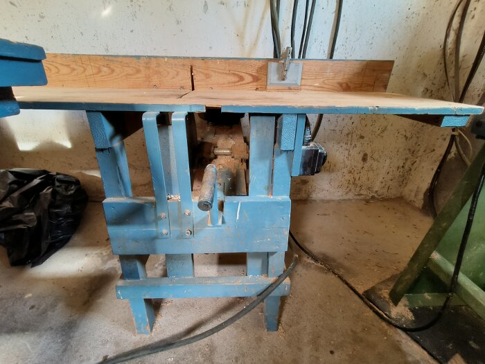 Blå träbearbetningsmaskin från 50-talet i garage, placerad på ett trästativ. Knappar och reglage synliga, elektriska kablar i bakgrunden.