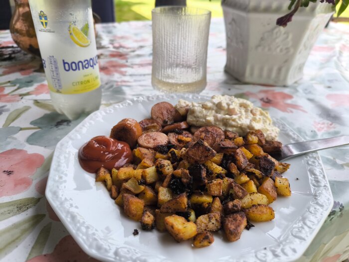 Bratwurst med råstekt potatis, lök, Dallassallad och ketchup på tallrik, med mineralvattenflaska och glas på blommig bordsduk.