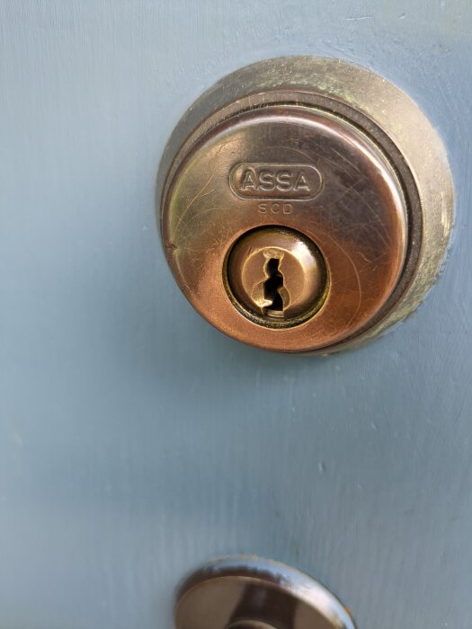 Låscylinder på en dörr av märket ASSA, inskjuten nyckel och sliten metall, problem med att låsa upp dörren utifrån.