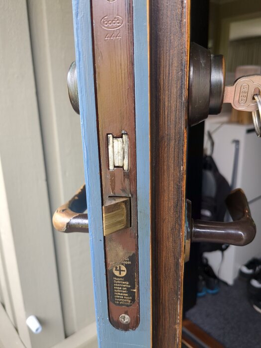 Närbild av ett dörrlås med nyckel i cylindern. Det är delvis synligt med text och ett handtag i trä intill.