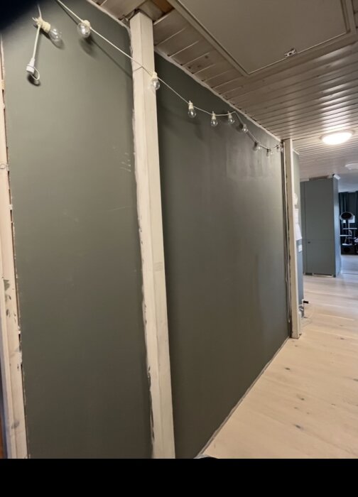 Grönmålad vägg under renovering med ljusslinga ovanför, omgiven av vit takpanel och trägolv.
