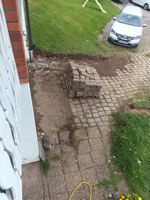 En hög med gatsten bredvid en uppriven trädgårdsgång av plattor intill ett hus, med gräsmatta och parkerad bil i bakgrunden.