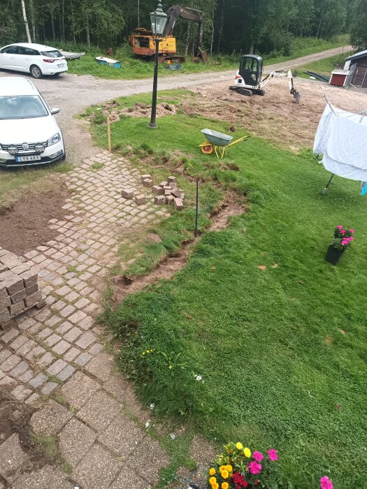 Grävarbete pågår i trädgård med maskin och stenläggning. En skottkärra står på gräset bredvid en hög med stenplattor.