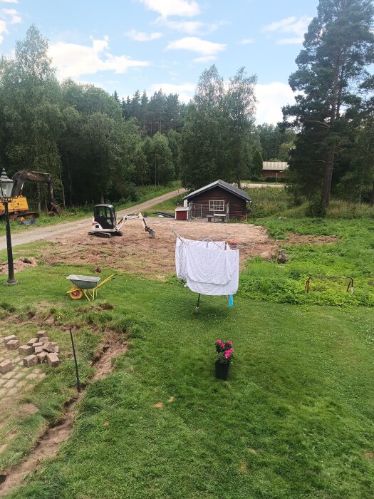 Grävmaskin vid grusyta framför ett rött hus, med grön gräsmatta, skottkärra och torkställning med kläder i förgrunden.