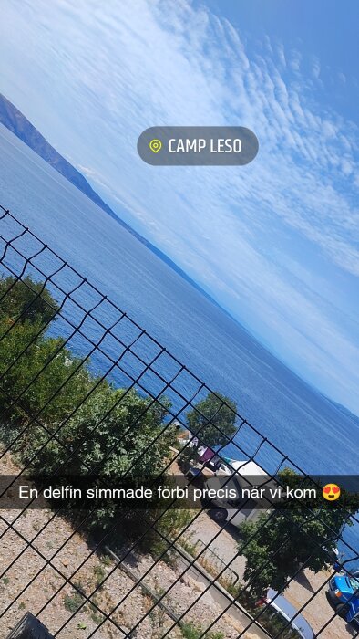 Utsikt över havet från Camp Leso med staket i förgrunden och bilar parkerade vid stranden, molnig himmel ovanför.