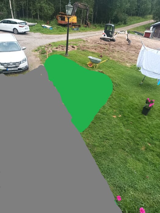 Gräsmatta med markering av framtida grusyta, grävmaskin i bakgrunden, skottkärra på gräset, och bilar parkerade på tomten.