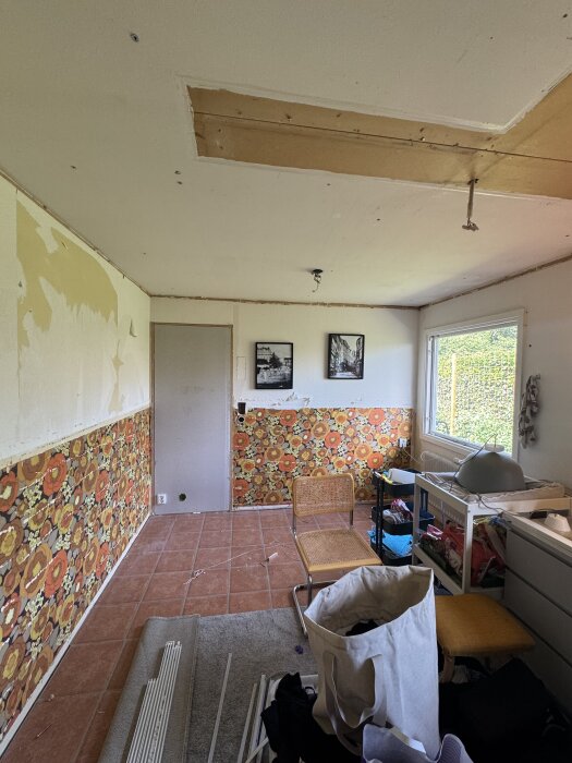 Rum under renovering med ett delvis borttaget innertak, retro blommig tapet, olika möbler och renoveringsmaterial på golvet.