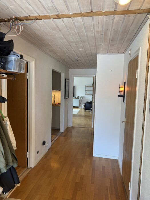Hall med trätak och synliga elkablar; dörrar till vänster och höger; golvet i trä; väggar vita; ljus i taket; öppning mot andra rum.