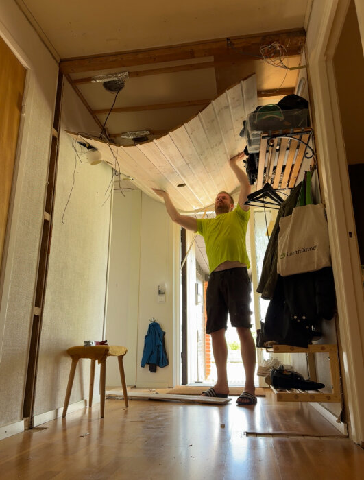 Person lyfter av innertak i hall, avslöjar eldosor och kablar. Golvet är täckt av verktyg och bråte. Renoveringsprojekt i bostadsmiljö.