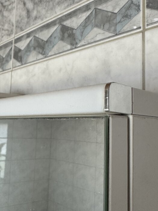 Vitt badrumsskåp med detaljbild på hörn och spegeldörr, kakelvägg med mönstrad fris i bakgrunden.