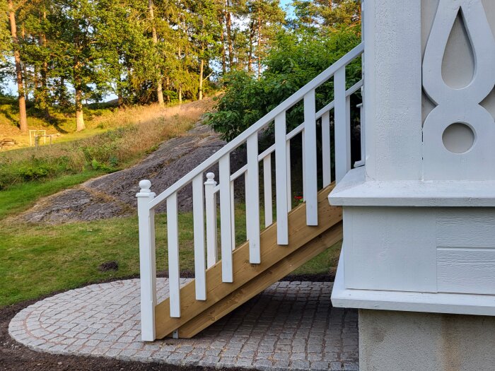 Vitmålat trappräcke med jämnt placerade ribbor, omgivet av en stenlagd yta och grönska i bakgrunden.