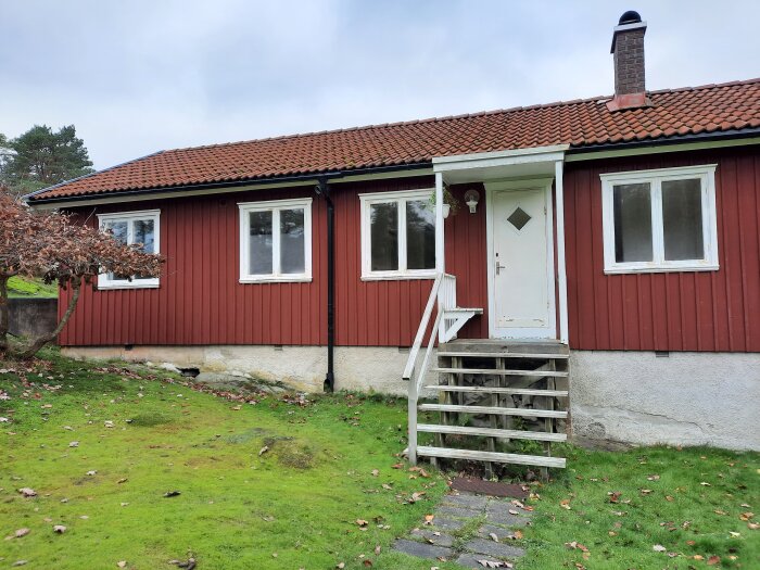 Rött hus med vita fönster och dörr, trappa upp till entrén, omgiven av grön gräsmatta och ett träd med höstlöv.