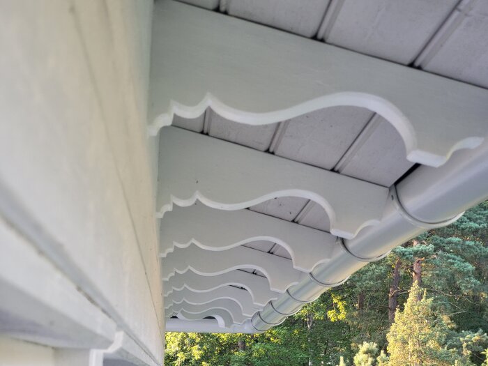 Dekorativa vita taksparrar med utsmyckningar under takfot på utomhusprojekt, mot bakgrund av grönska.