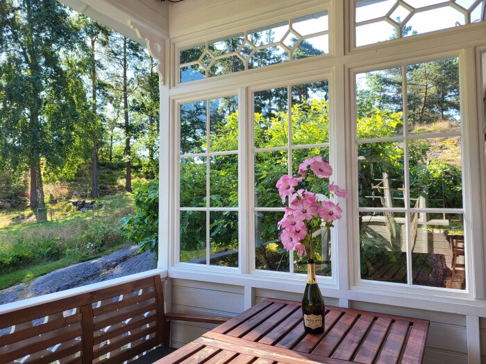 Bilden visar ett uterum med träbord, rosa blommor i en champagneflaska och grönskande trädgård utanför fönstren.
