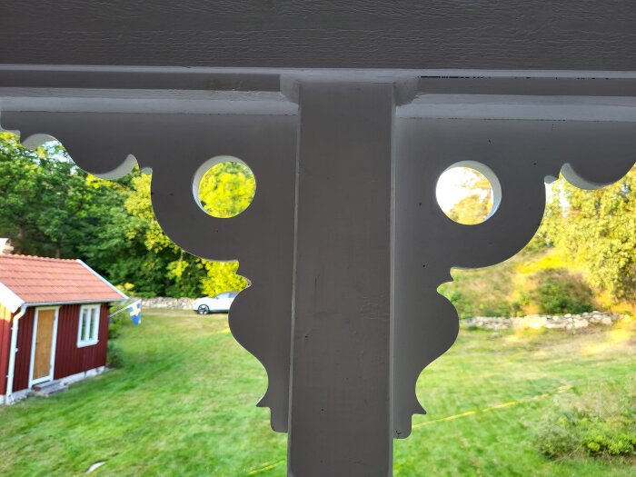 Dekorativa vita trädetaljer med utsmyckningar, sett mot en grönskande bakgrund och ett rött hus med tegeltak i ett landskap.