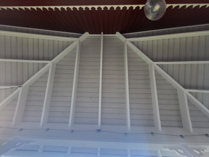 Takdetalj i trä med vita brädor och lamprad, röd panel upptill och en del av en glaslampa synlig.