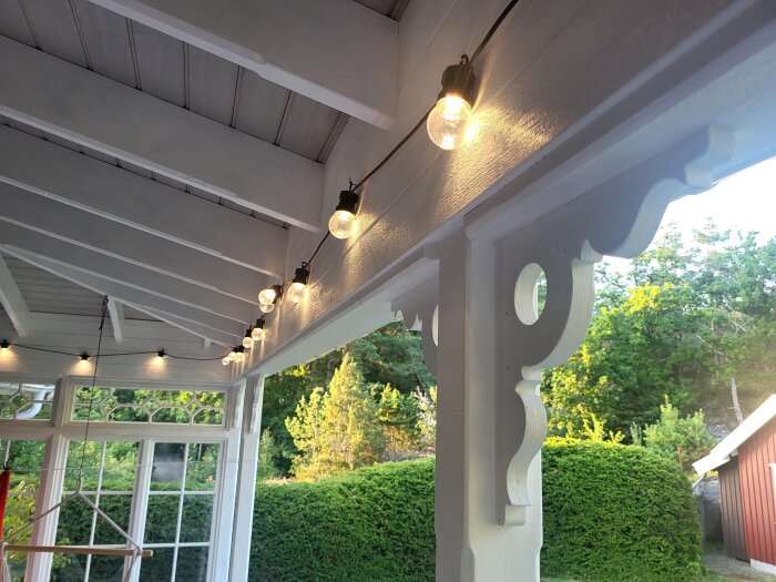 Upphängda glödlampor under ett vitmålat verandatak med dekorativa trädetaljer och fönster, omgivna av grönska i bakgrunden.