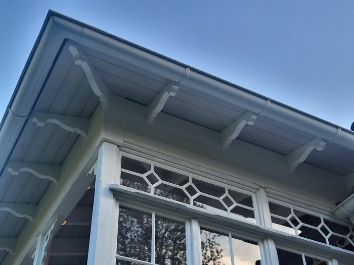 Dekorativt snickarglädje under taket på en vitmålad veranda med detaljerade fönster.