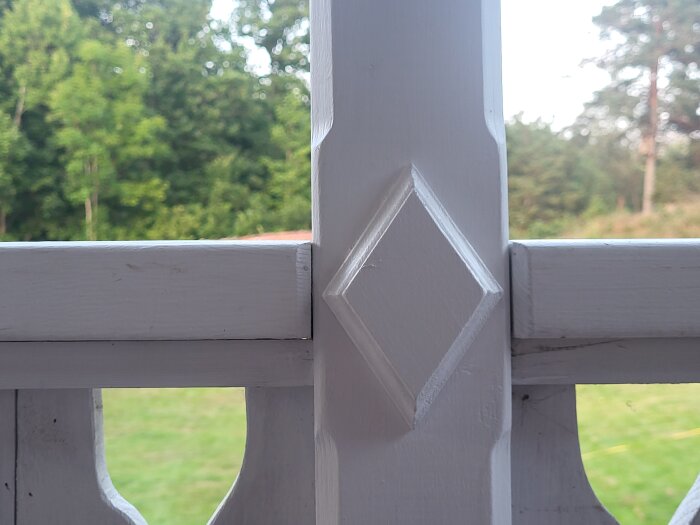 Detalj av vitmålat räcke med en diamantformad dekoration, omgiven av grön bakgrund utomhus.