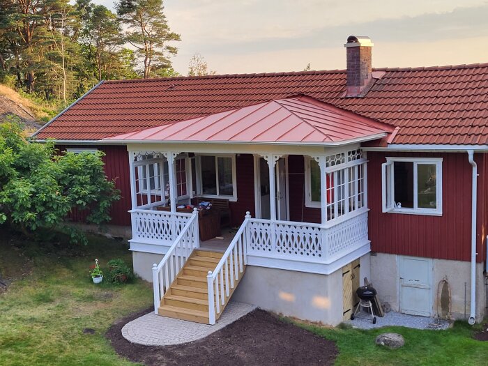 Rött hus med nytt vitt träveranda och trappa, omgiven av grönska under en molnfri himmel. Chimenea synlig på taket.