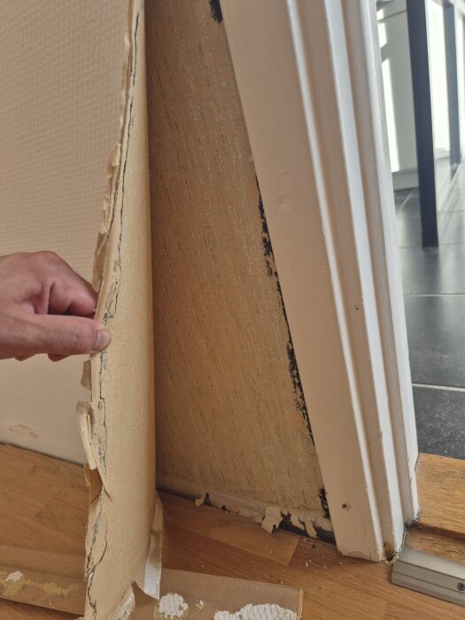 En person drar bort gammal tapet från en vägg, exponerar en äldre tapet under. Golvlister och golv syns.