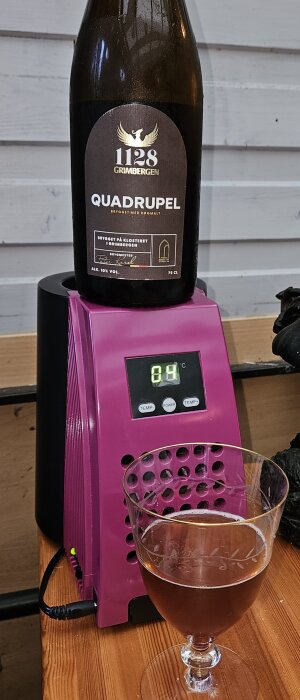 Grimbergen Quadrupel-flaska ovanpå en rosa ölkylare, med ett glas öl framför på ett träbord.