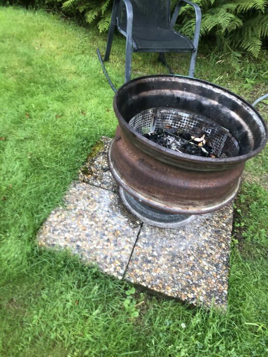 Eldplats byggd av bil- och lastbilsfälgar, placerad på stentplattor i en grönskande trädgård med en stol i bakgrunden.