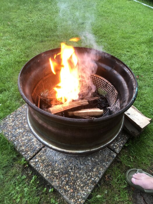 Eldstad av bil- och lastbilsfälgar på trädgårdsplattor, brinnande ved och grillgaller inuti, omgiven av gräs, fot synlig vid kanten.