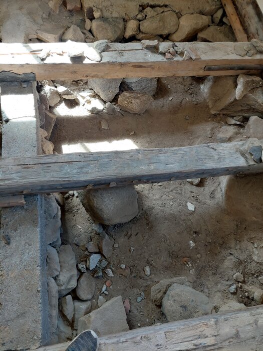 Träbjälklag med synliga stödmurar och ojämn jord på marknivå, stora stenar och smuts framtill. En fot syns i nedre vänstra hörnet.