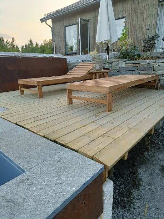 Utomhusmiljö med trädäck, två solsängar, ett parasoll, planteringar och del av skifferkant vid ett hus.