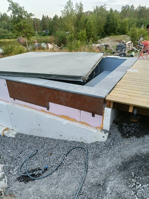 Skifferkantat poolområde under konstruktion, täckt med presenning, bredvid en träveranda och omgivet av gröna träd och en grusgång.