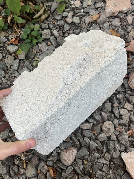 En hand håller en ljusgrå betongblockbit mot en bakgrund av grus och småstenar.