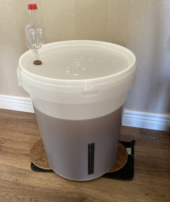 Plastbehållare med ölbryggning på gång, utrustad med jäsrör, står på ett trägolv.