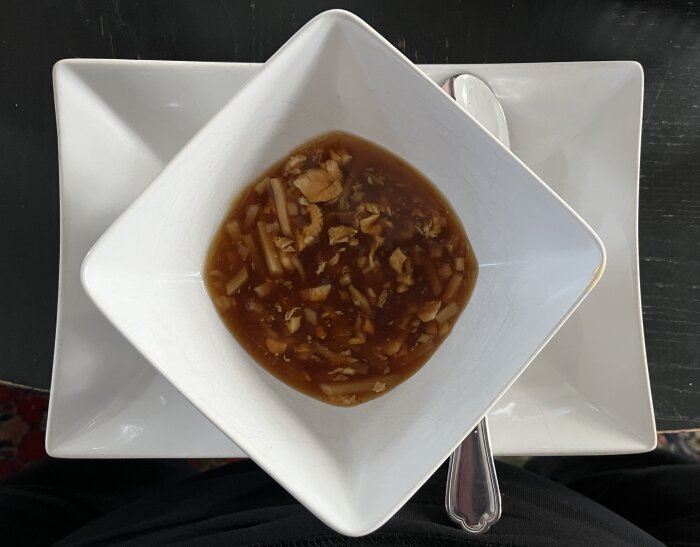 Vitt fyrkantigt fat med skål fylld med Peking-soppa, synliga grönsaksbitar i brun buljong.