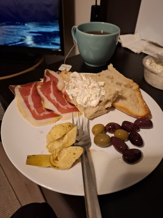 Tallrik med parmaskinka, bröd med röra, oliver och kronärtskockor. En kopp te och TV i bakgrunden.