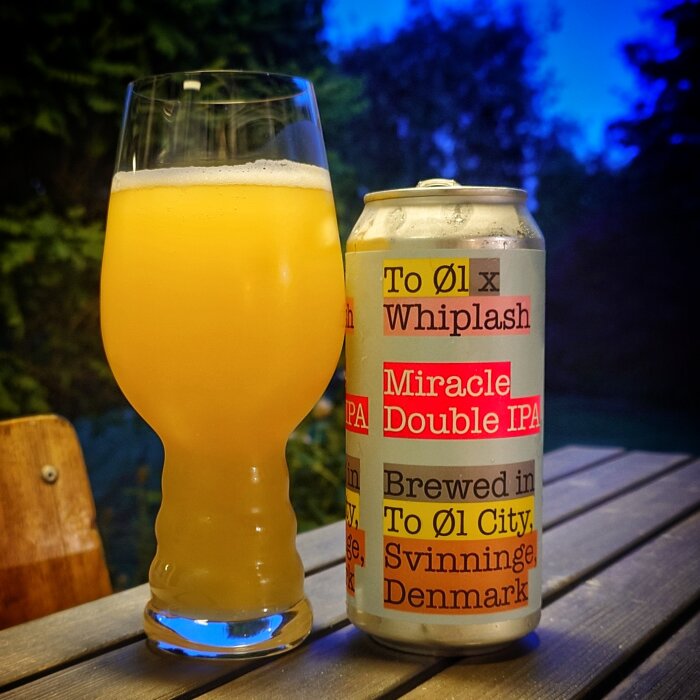 Ett glas med gyllene Double IPA-öl bredvid en burk med texten "Miracle Double IPA" i en utomhusmiljö.