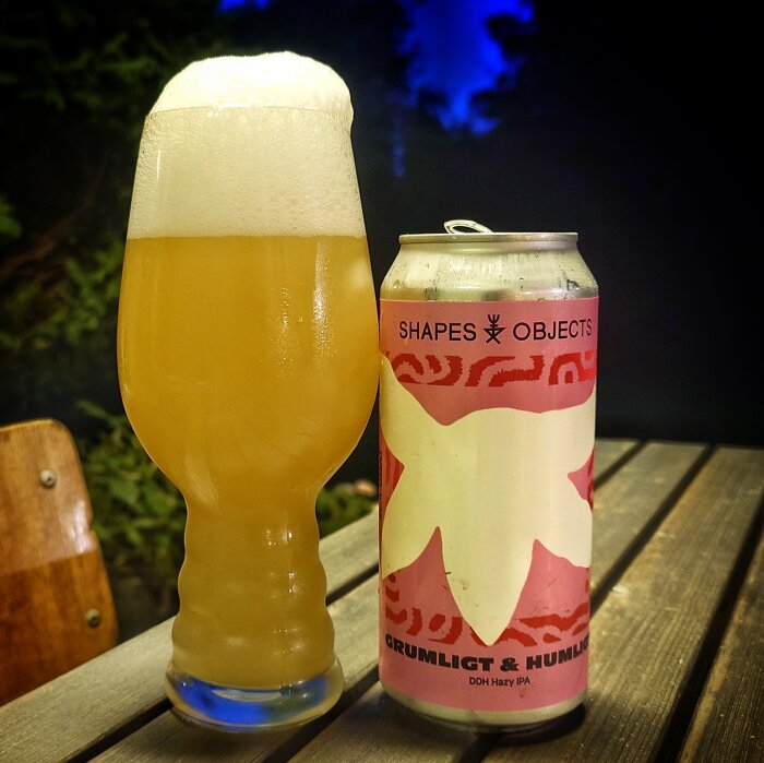 En ölburk med etiketten "Shapes Objects" och "Grumligt & Humligt" står bredvid ett glas skummande öl på ett trädäck på kvällstid.