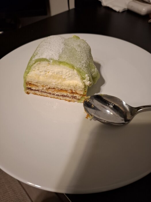 Prinsesstårta på en vit tallrik med en sked, biten är täckt med grönt marsipanöverdrag och har flera lager inuti.