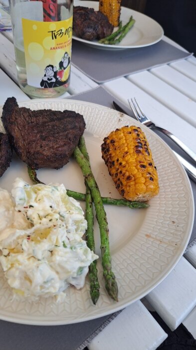 Tallrik med grillat lamm och picanha, majskolv, sparris och potatissallad. En flaska cider syns i bakgrunden.