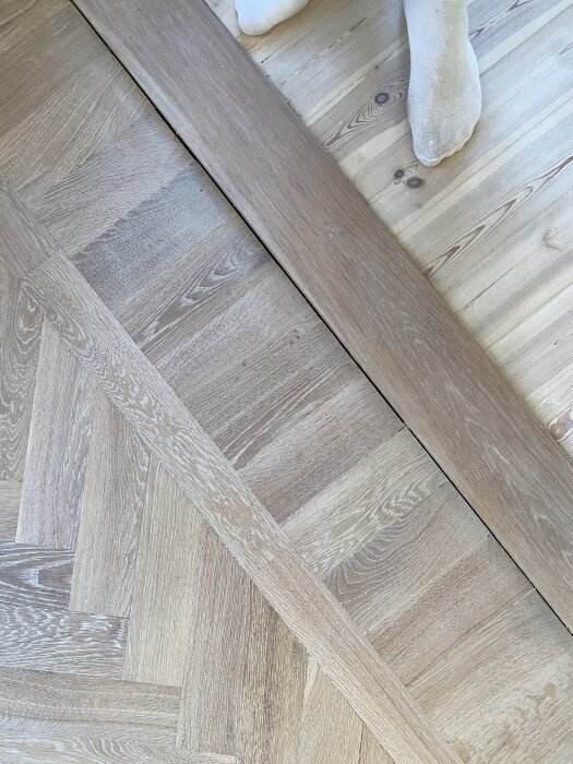 Detaljbild av ett äldre ekparkettgolv och furutiljor som visar färgskillnader, repor och defekt fogning samt fot och strumpa i bakgrunden.