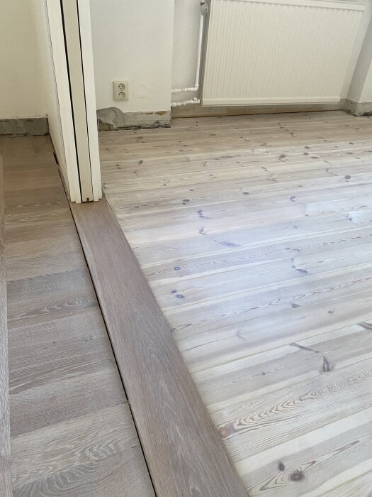 Trägolv med övergång mellan två olika typer av trä, ljus furugolv och mörkare ekplank, intill en vägg med radiator och eluttag.