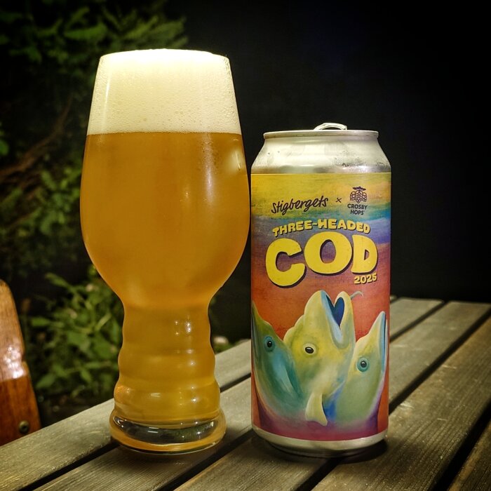 En ölburk med etiketten "Three-Headed Cod 2023" står bredvid ett glas öl på ett träbord utomhus.