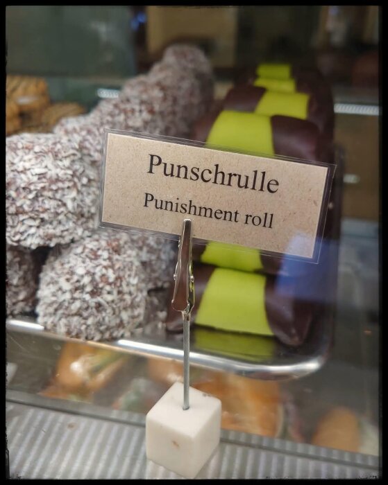Punschrullar med choklad och grön marsipan visas i ett konditori, med en handskriven skylt som felaktigt översatt till "Punishment roll".