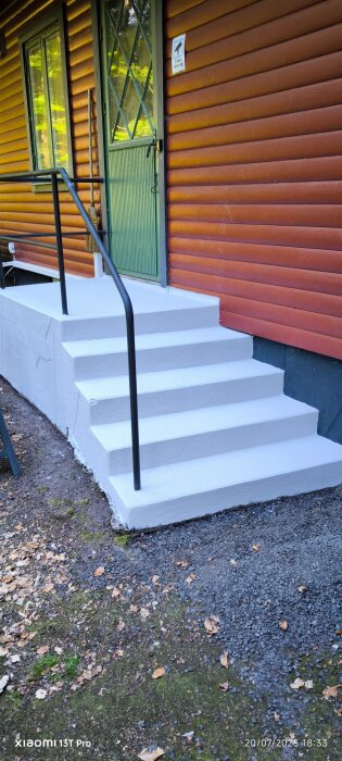 Vitmålad betongtrappa med svart räcke vid ett rödbrunt trähus med grön dörr, omgiven av grus och mossa.