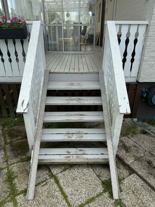 Vit trappa med räcken, belägen vid ett hus med inglasad veranda. Trappan står på en stenlagd yta och visar tecken på slitage.
