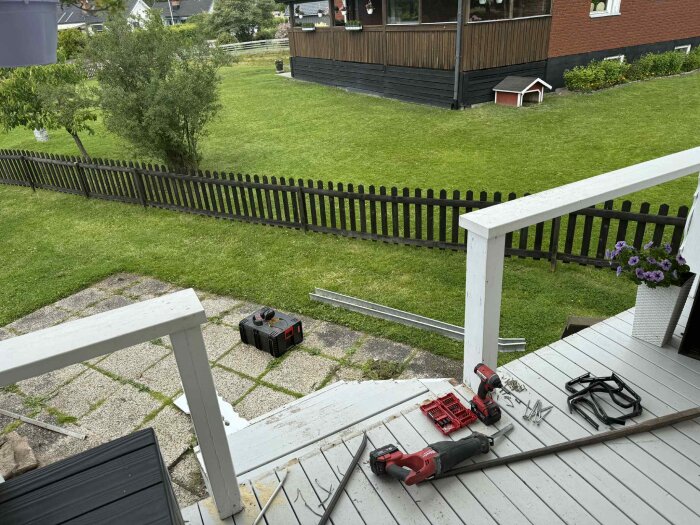 Verktyg och material för renovering på en altan; synliga trappsteg och staket, gräsmatta och verktygslåda i bakgrunden.