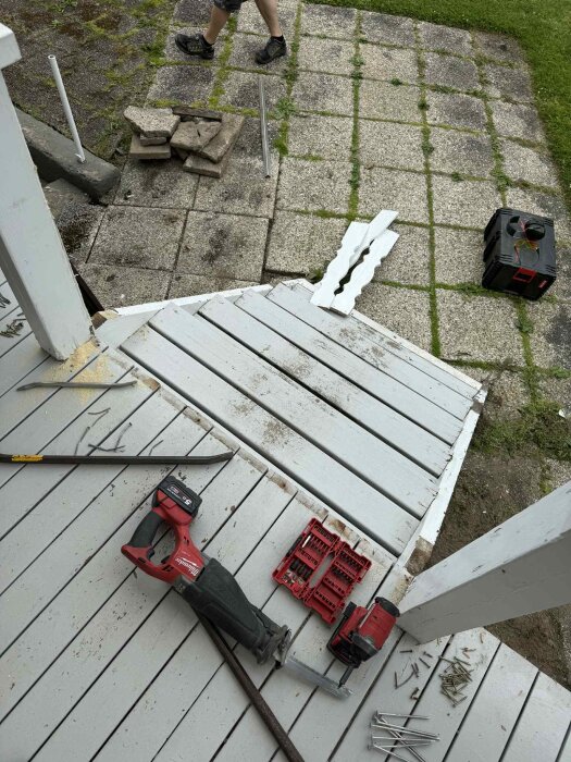 Verktyg och material på en veranda redo för renovering, inklusive en såg, skruvar och lösa brädor vid en rivningsplats nära trappan.